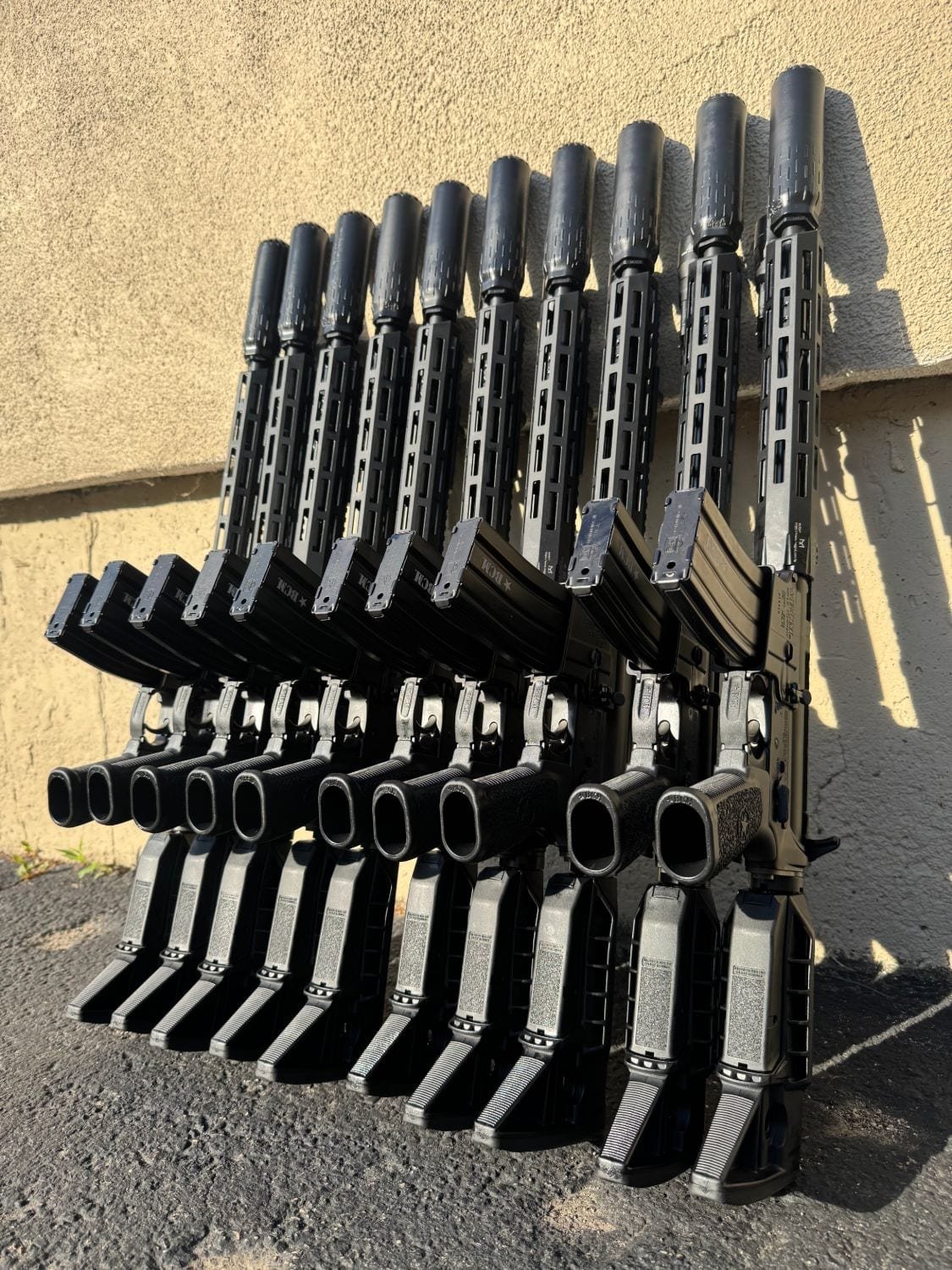 Row of suppressed BCM rifles leaning against a sunlit wall