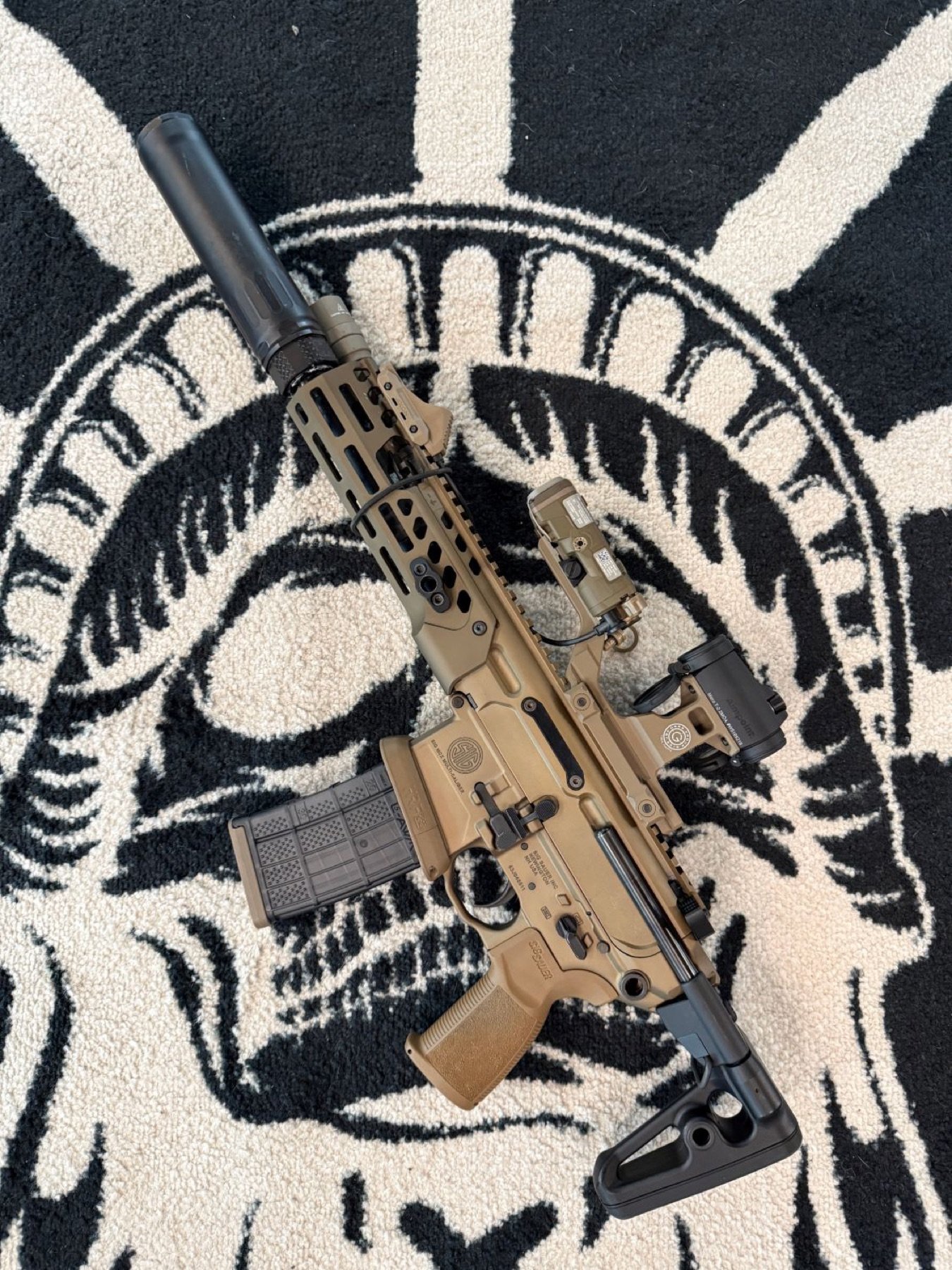 A suppressed SIG Sauer rifle with optic and light on the Liberty Ordnance Supply logo rug